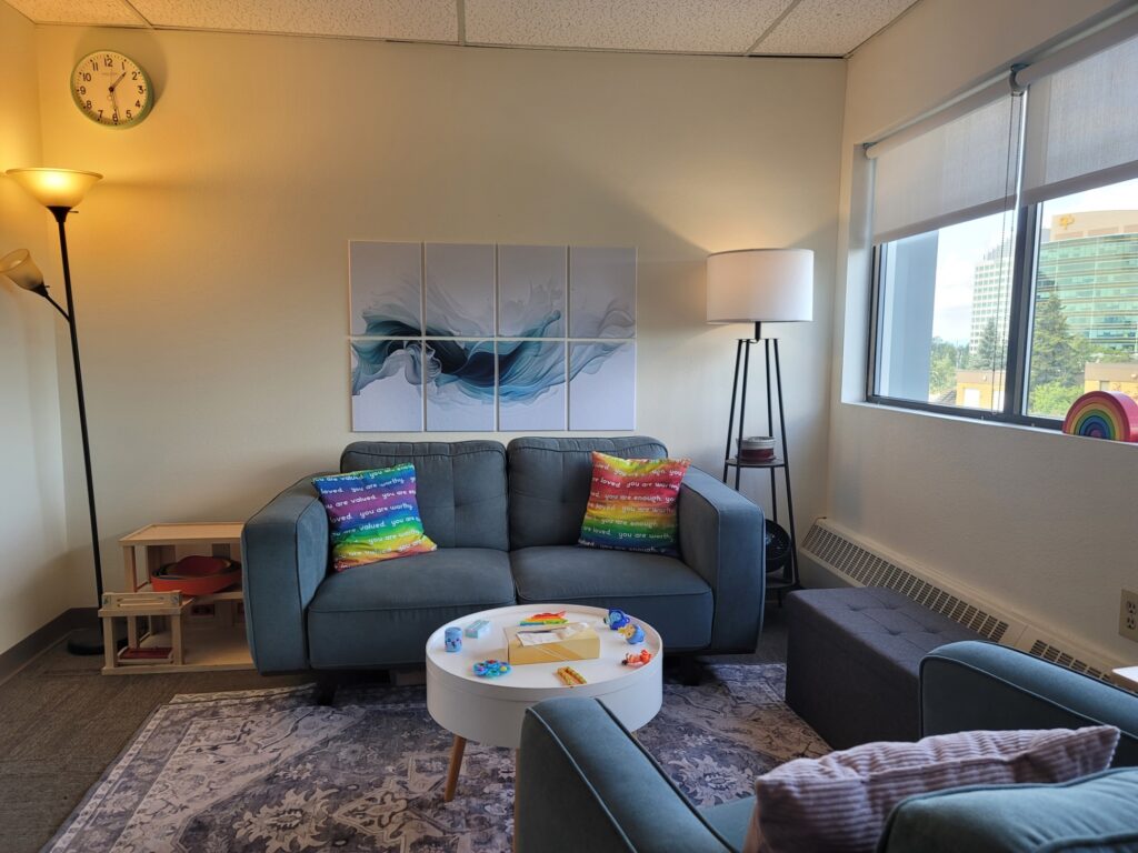 View of Playful Connection Counseling Family Couch with rainbow pillows that say "you are enough". Behind the Couch is a fluid felt tile image, lamps are on each side of the couch, a round table is in front of the couch and a storage ottoman is located next to the window for additional seating