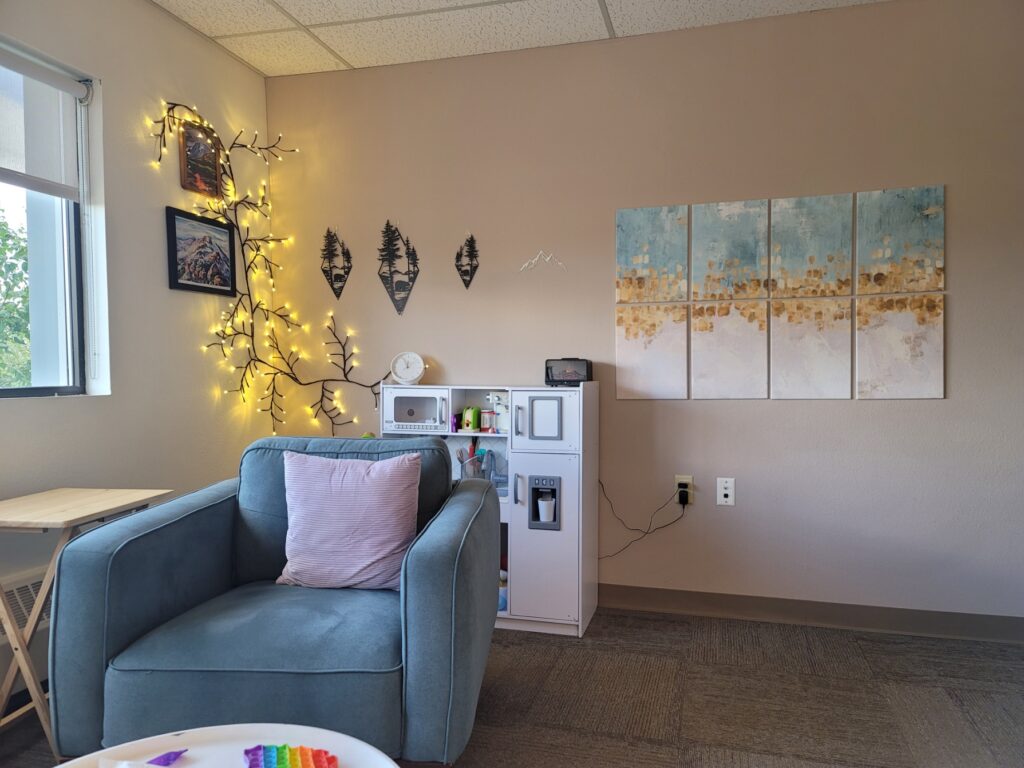 Client view at Playful Connection Counseling. A chair with pillow is located in the front, with a child's play kitchen located behind the chair. On the wall is a vine of lights, metal mountain art, and another felt tile artwork.