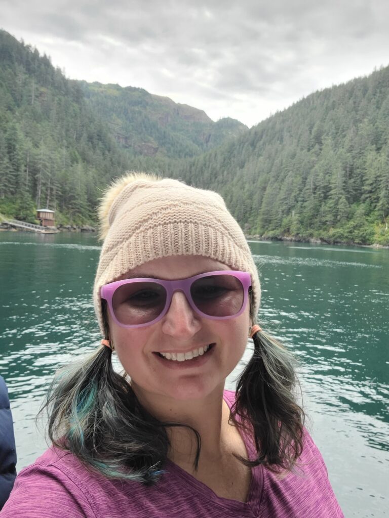 Image of Brandi Askeland of Playful Connection Counseling smiling into the camera, wearing glasses and a beanie hat. The background is mountains and water of Resurrection Bay, Alaska. In the distance a cabin can be seen over Brandi's shoulder.